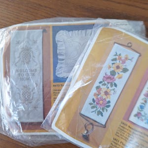 May include: Two embroidery kits with white fabric and colourful thread. The first kit features a welcome sign with a pineapple design. The second kit features a floral design with pink, yellow, and green flowers.