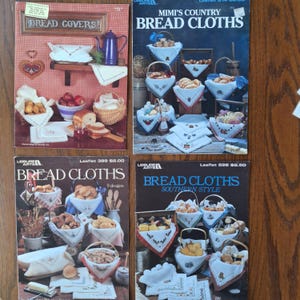 May include: Four vintage craft leaflets featuring bread cloths and covers. The covers are displayed with bread, fruit, and baskets. The leaflets include titles such as "Bread Covers", "Mimi's Country Bread Cloths", and "Bread Cloths Southern Style".