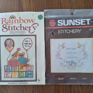 Könnte beinhalten: Zwei Vintage-Kreuzstich-Kits. Das "Rainbow Stitchery"-Kit zeigt ein Baby-Ankündigungsdesign mit einem Clown, einem Teddybären und Alphabetblöcken. Das "Sunset Stitchery"-Kit ist ein süßes Baby-Andenken mit einem floralen Kranzdesign.