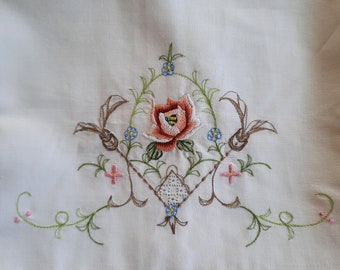 May include: White embroidered pillowcase with a floral design featuring a pink rose with a yellow center, surrounded by green leaves and small blue flowers. The pillowcase has a yellow trim.