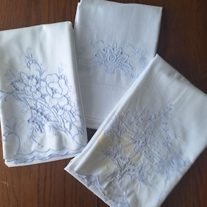 May include: Three white rectangular linens featuring intricate light blue floral embroidery. Each piece displays a unique pattern of flowers and leaves, with scalloped edges on two of the items and detailed cutwork designs throughout the fabric.