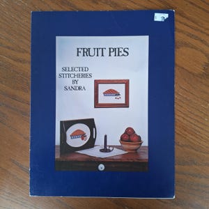 Può includere: Una copertina di libro intitolata "Fruit Pies" con il sottotitolo "Selected Stitcheries by Sandra". La copertina presenta illustrazioni a punto croce di torte alla frutta, una ciotola di mele e un portacandele su una superficie di legno.