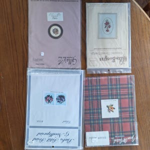 May include: Four cross-stitch or needlepoint kits are displayed. The kits feature floral designs, with one in a circular frame and another in an oval frame. The kits are packaged in plastic and paper, with text labels and brand names visible.