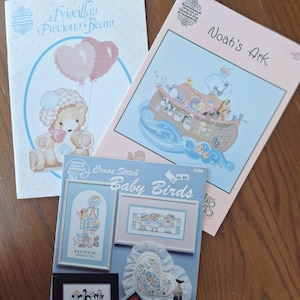 Peut inclure: Trois modèles de point de croix représentant un ourson avec des ballons, l'arche de Noé et des bébés oiseaux. Les modèles sont imprimés sur du papier rose et bleu clair avec une bordure blanche.