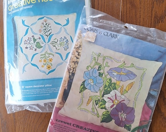 May include: Two embroidery kits for pillows. The first kit features a white pillow with blue and yellow floral embroidery. The second kit features a white pillow with blue and purple morning glory embroidery. Both kits include instructions and embroidery floss.
