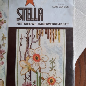 Peut inclure: Un kit de point de croix avec un motif floral de jonquilles et un logo d'étoile rouge. Le kit est intitulé "Stella Het Nieuwe Handwerkpakket" et comprend le texte "Spring Time, Artikel Nr. 1064, Made in Holland, Ontwerp: Lore Van Dijk".