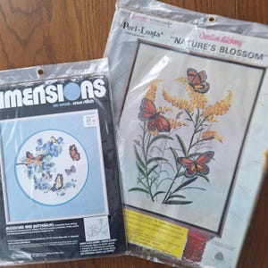 Peut inclure: Deux kits de point de croix. L'un présente un motif floral bleu avec des papillons et le texte "BLOSSOMS AND BUTTERFLIES". L'autre kit est intitulé "NATURE'S BLOSSOM" avec des papillons et des fleurs jaunes. Les deux kits sont dans un emballage plastique.