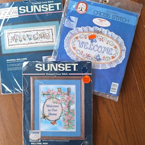 May include: Three cross stitch kits featuring welcome signs. The first kit is a white background with a blue welcome sign with a floral design. The second kit is a white background with a blue welcome sign with a floral design and a white lace border. The third kit is a blue background with a welcome sign with a floral design and a butterfly.