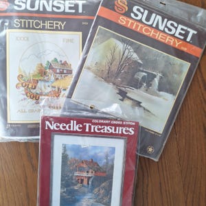 May include: Three cross-stitch kits. Two are Sunset Stitchery brand, one with a winter scene and the other with a mill. The third kit is Needle Treasures brand, featuring a covered bridge scene. All kits are in plastic packaging.