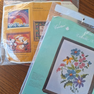 May include: Two cross-stitch kits in plastic packaging. One kit features a rainbow design, the other a floral spring garden. The rainbow kit is 8 x 10 inches (20.3 cm x 25.6 cm) when finished. The spring garden kit is 11 x 14 inches (27 cm x 35 cm).