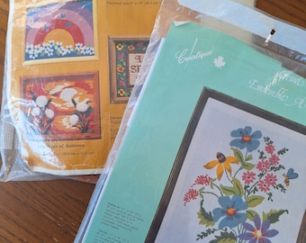 May include: Two cross-stitch kits in plastic packaging. One kit features a rainbow design, the other a floral spring garden. The rainbow kit is 20.3 cm x 25.6 cm when finished. The spring garden kit is 27 cm x 35 cm.