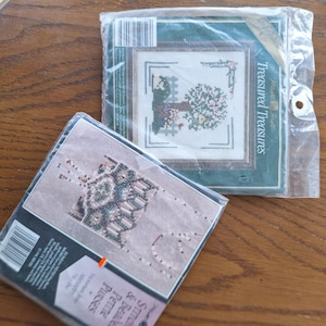 May include: Two sealed craft kits in plastic packaging. One kit is titled Treasured Treasures and features a cross-stitch pattern of a tree and fence. The other kit is titled Victorian Beaded Petite Purses and displays a beaded purse design.
