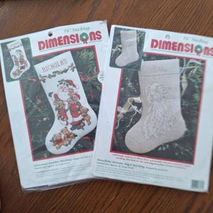 May include: Two Dimensions brand 16-inch Christmas stocking counted cross-stitch kits in plastic packaging. One kit features a Santa Claus with teddy bears and the name Nicholas, while the other shows an angel design with the name Ashley.