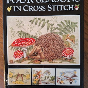 May include: Book cover for "Four Seasons in Cross Stitch" featuring a hedgehog surrounded by mushrooms and ferns. The cover also includes three smaller images of cross stitch patterns: a rabbit, a dragonfly, and bluebirds at a bird feeder.