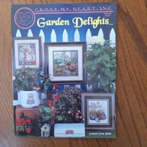 May include: The cover of a magazine titled "Garden Delights" showcasing framed cross-stitch art depicting garden scenes. The cover includes images of flowers, plants, and a small house. The text "Cross My Heart" is also visible.