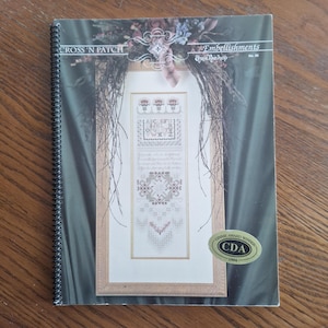 May include: A spiral-bound book titled "Cross 'N Patch" featuring a framed cross-stitch design. The design includes floral elements, an alphabet, and geometric patterns. The cover also displays the text "Embellishments" and a "Ginnie Award Winner" sticker.