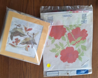 May include: Two needlepoint kits in clear plastic packaging. One kit features a bird design with brown and tan birds on a grid canvas. The other kit displays a floral pattern with three large red flowers and green leaves on a white grid canvas.