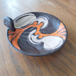 May include: A small, round ceramic dish featuring a dark charcoal gray base color. The surface is decorated with abstract, flowing patterns in bright orange and creamy white. A small, dark gray handle protrudes from one side of the dish rim.