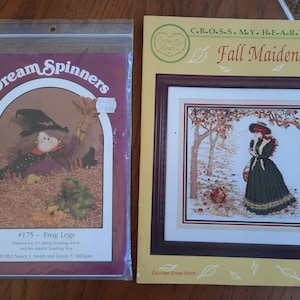 May include: Two cross stitch patterns. The first pattern is for a witch doll with a frog. The second pattern is for a woman in a green dress walking through a forest with red leaves. The text on the second pattern reads "Cross My Heart, Fall Maiden".
