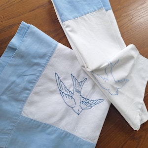 May include: Two white pillowcases with blue embroidered bird designs. The pillowcases have a light blue border. One pillowcase is folded, and the other is unfolded, displaying the embroidered bird.