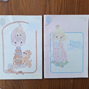 Puede incluir: Dos patrones de punto de cruz Precious Moments. Uno presenta a un niño con un perro y un oso, el otro a una niña con un cordero y un perro. Ambos tienen las palabras "Precious Moments" en cursiva.