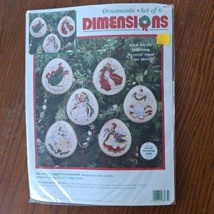 Può includere: Un kit per punto croce contato del marchio Dimensions per sei ornamenti angelo di forma ovale. La confezione riporta il testo Mystical Angel Ornaments, Set of 6, e indica una dimensione finita di circa 11 cm di altezza per ogni ornamento.