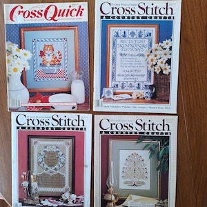 May include: Four cross stitch magazines with colorful covers featuring various designs. The covers include a cat, a sampler with the alphabet and numbers, a floral design, and a family tree.