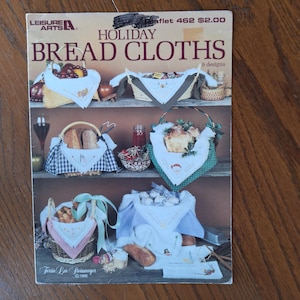 Può includere: Un libretto vintage intitolato "Holiday Bread Cloths" di Leisure Arts, con nove disegni. La copertina mostra vari cestini di pane con tovaglie decorative, che mostrano temi festivi e ricami. Il libretto è su una superficie di legno.
