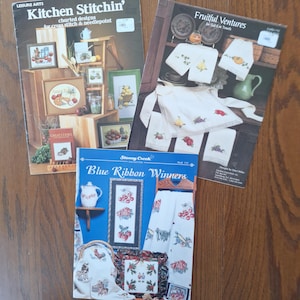 Può includere: Tre libri di modelli a punto croce sono esposti su una superficie di legno. I libri si intitolano "Kitchen Stitchin'," "Fruitful Ventures" e "Blue Ribbon Winners." Le copertine presentano immagini di disegni a punto croce, tra cui motivi a tema cucina e motivi floreali.