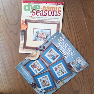 Può includere: Due libri di modelli a punto croce. Il libro in alto si intitola "dye-namic Seasons" con un disegno floreale a punto croce. Il libro in basso si intitola "SEASONAL SCENES" con quattro disegni a punto croce incorniciati. I libri sono su una superficie di legno.