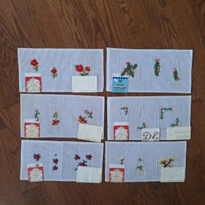 May include: Eight white canvas panels with a grid pattern, each featuring a different embroidered design. The designs include flowers, butterflies, ladybugs, and holly. Each panel has a small white label with the text "Luxury Needlepoint" printed on it.