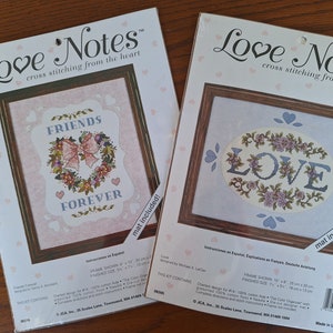 Puede incluir: Dos kits de punto de cruz Love Notes, uno con un fondo rosa y las palabras "Friends Forever" en una corona, y el otro con un fondo azul y la palabra "LOVE" en un óvalo floral. Ambos kits incluyen una alfombra.