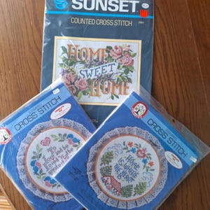 May include: Three cross-stitch kits. One kit features the text "HOME SWEET HOME" surrounded by roses. Two kits have circular designs with the text "To love and be loved is the greatest" and "Home is where the heart is".
