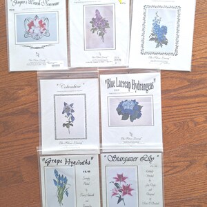 Könnte beinhalten: Sieben Kreuzstich-Sets in Kunststoffverpackungen werden gezeigt. Die Sets zeigen florale Designs, darunter "Ginger's Beach Nouveau", "Lavender Lilacs" und "Delphiniums". Jedes Set enthält ein gerahmtes Blumenbild und Text. Die Sets liegen auf einer Holzoberfläche.