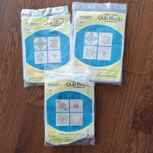 Puede incluir: Tres paquetes de bloques de edredón WonderArt. Cada paquete presenta un diseño azul y amarillo con varios patrones de bordado floral y geométrico. Los paquetes están sellados en plástico transparente.