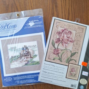 May include: Two counted cross stitch kits in plastic packaging. The left kit, An Amish Afternoon, shows a horse-drawn carriage on a path. The right kit, Graceful Tulip, features a large pink tulip flower with green leaves on a beige background.
