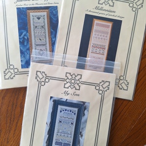 May include: Three cross-stitch patterns, each with a different design. The first pattern is titled "Jasmine and Sapphires" and features a blue background with a floral design. The second pattern is titled "Millennium" and features a white background with a geometric design. The third pattern is titled "My Son" and features a blue background with a floral design.
