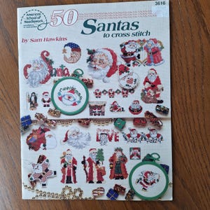 May include: A cross stitch pattern booklet featuring 50 different Santa Claus designs. The cover features a colorful illustration of Santa Claus with a red hat and beard, surrounded by other festive designs. The title of the booklet is "50 Santas to cross stitch" by Sam Hawkins.