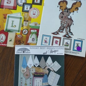 Può includere: Tre libri di modelli di punto croce. Il primo libro è giallo con un tema alfabeto da cucina. Il secondo libro è bianco con un alce e il titolo "Kouge Kappie Chocolate Mousse and Other Kitchen Capers". Il terzo libro è marrone con un titolo che recita "Sunday Best Breadcovers and Recipes".
