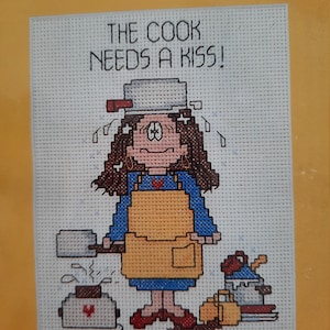Puede incluir: Patrón de punto de cruz con una mujer de dibujos animados que lleva un vestido azul y un delantal amarillo, sosteniendo una espátula y una olla. El texto "THE COOK NEEDS A KISS!" está por encima de la mujer.