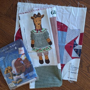 Könnte beinhalten: Ein Bastelset mit einer Anleitung für eine Giraffenpuppe und eine "Puppy Love"-Puppe. Das Set enthält Stoff, Anleitungen und Stickgarn. Die Giraffenpuppe trägt ein grün-weißes Kleid mit roten Akzenten. Die "Puppy Love"-Puppe trägt ein rot-weißes Kleid mit einem Herz auf der Vorderseite. Das Set ist in einer durchsichtigen Plastiktüte mit einem rot-weißen Etikett verpackt.