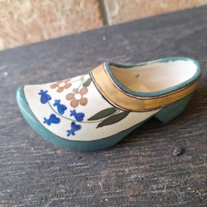 May include: A ceramic clog, painted with a floral design in brown and blue on a cream background. The rim is trimmed with gold and green. The shoe is resting on a dark wooden surface.