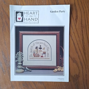 Puede incluir: Un patrón de bordado impreso titulado "Garden Party" de Heart in Hand Needleart. El diseño presenta un punto de cruz enmarcado con el texto "In search of the perfect flowers". La imagen incluye un objeto decorativo de metal y flores secas.