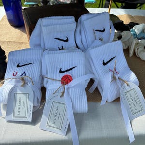 May include: Several pairs of white crew socks featuring black embroidered swoosh logos and small graphic icons including a basketball, a baseball, and the text USA. Each pair is tied with a white ribbon and a tag labeled LITTLELALOOS.
