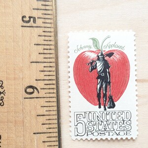 May include: A vintage 5-cent United States postage stamp featuring a red apple with a green stem and leaves. The apple is split in half to reveal a black and white illustration of Johnny Appleseed holding a shovel and a bag. The text "Johnny Appleseed" is written above the apple and "United States Postage" is written below the apple.
