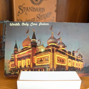 Könnte beinhalten: Vintage Postkarte mit dem World's Only Corn Palace in Mitchell, South Dakota. Die Postkarte zeigt ein hell beleuchtetes Gebäude mit mehreren Kuppeln und Flaggen, mit dem Text "World's Only Corn Palace".