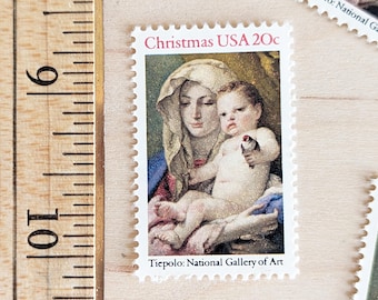Pode incluir: Um selo postal de Natal vintage que apresenta uma pintura da Madona e o Menino. O selo é branco com uma borda vermelha e o texto "Christmas USA 20c" em vermelho. A pintura representa uma mulher com roupas azuis e brancas segurando uma criança pequena. O texto "Tiepolo: National Gallery of Art" está impresso abaixo da pintura.