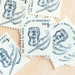 May include: A collection of vintage USA 1¢ stamps. Each stamp features a quill pen, inkwell, and the text "The Ability to Write - A Root of Democracy." The stamps are white with blue ink and have a scalloped edge.