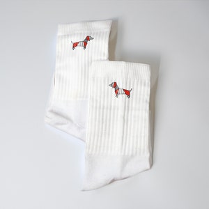May include: Two pairs of white socks featuring a dachshund design. The dachshund is wearing a red and white striped jumper.