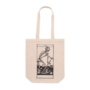 Può includere: Borsa tote in tela beige con un'illustrazione in bianco e nero di una figura di scheletro con una falce, incorniciata da un bordo rettangolare. Il numero romano XIII è in alto. La borsa ha due manici.